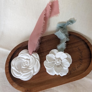 Könnte beinhalten: Zwei weiße Keramik-Blumenornamente mit einem zarten Design, eines in Rosenform und das andere in einer offeneren Blütenform. Die Ornamente werden auf einem Holztablett mit dem eingravierten Text "gibt ein bisschen" präsentiert. Die Ornamente sind mit rosa und blauen Bändern gebunden.