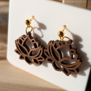 Könnte beinhalten: Ein Paar Holzohrringe mit einem Lotusblumen-Design. Die Ohrringe sind braun und haben goldfarbene Haken.