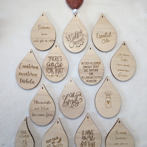 Könnte beinhalten: Eine Sammlung von 12 Holz-Tropfen-Anhängern mit verschiedenen Sprüchen über ätherische Öle. Die Anhänger sind mit Lasergravur versehen mit Texten wie "Y'all Need Oils", "There's an Oil for That", "Essential Oils", "Oil Lover", "Keep Calm and Clary On", "I Have an Oil for That", und "In den kleinen Dingen zeigt die Natur die allergrößten Wunder".