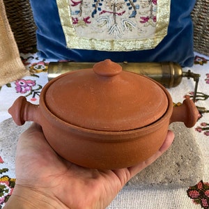 Puede incluir: Una olla de cocción de terracota con dos asas y una tapa. La olla es de color marrón y tiene un aspecto rústico.