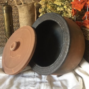 Könnte beinhalten: Ein Terrakotta-Topf mit Deckel, der Deckel ist leicht geöffnet und gibt den Blick auf das schwarze Innere des Topfes frei. Der Topf steht auf einem weißen Tuch, im Hintergrund eine in Sackleinen verpackte Flasche.