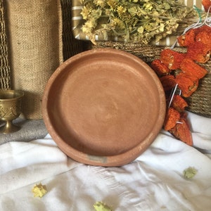 May include: A terracotta plate sits on a white cloth, with dried herbs and red peppers in the background. A bottle in a burlap sack and a small brass cup are also visible. The plate is round and has a slightly raised edge.