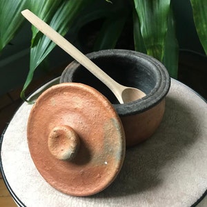 May include: A small, terracotta pot with a lid and a wooden spoon inside. The pot has a dark brown glaze on the inside and a reddish-brown glaze on the outside. The lid has a small knob on top.