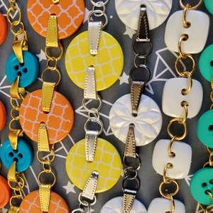 May include: A close-up of a colorful necklace made with various buttons and metal chain links. The buttons are in shades of orange, yellow, blue, green, and white. Some buttons have a geometric pattern, while others are solid colors.