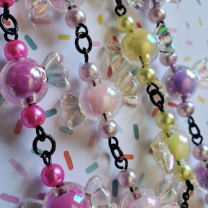May include: Close-up of beaded necklaces with candy-shaped beads in shades of pink, purple, and yellow. The beads have iridescent, clear wings and are strung on black chain. The background is a colorful, speckled pattern.