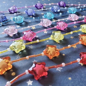 Könnte beinhalten: Nahaufnahme mehrerer Perlenstränge mit sternförmigen Anhängern in verschiedenen Farben, darunter Lila, Blau, Rosa, Gelb und Orange. Die Perlen sind auf dünnen, transparenten Schnüren aufgefädelt. Der Hintergrund ist eine dunkelblaue Oberfläche mit kleinen weißen Sternmustern.