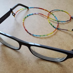 Könnte beinhalten: Schwarze Brille mit einer Regenbogen-Perlen-Brillenkette. Die Kette besteht aus kleinen, farbigen Perlen in einem Farbverlaufsmuster. Die Brille hat einen rechteckigen Rahmen und klare Gläser.