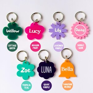 May include: A collection of colorful pet ID tags in various shapes, including a four-leaf clover, flower, and bell. Each tag features a pet's name in white lettering and a coordinating color, such as green, pink, purple, and orange. The tags are attached to silver key rings.