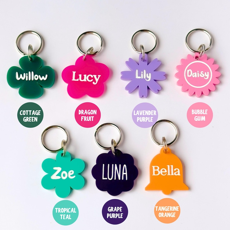 May include: A collection of colorful pet ID tags in various shapes, including a four-leaf clover, flower, and bell. Each tag features a pet's name in white lettering and a coordinating color, such as green, pink, purple, and orange. The tags are attached to silver key rings.