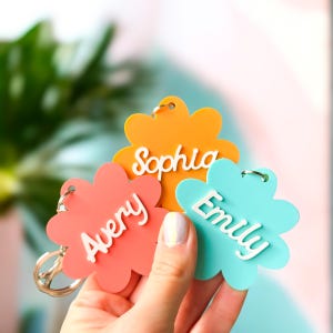 May include: Three flower-shaped keychains in coral, orange, and turquoise. Each keychain has a different name in white script: Avery, Sophia, and Emily. The keychains are held in a person's hand, with a blurred background.