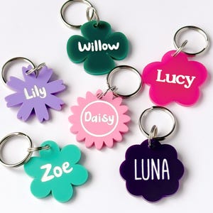 May include: A collection of colorful, flower-shaped keychains with silver key rings. Each keychain features a different name in white lettering: Willow, Lily, Daisy, Zoe, Lucy, and Luna. The keychains are in shades of green, purple, pink, and teal.