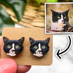 May include: A pair of wooden earrings featuring a black and white cat face design. The earrings are shown on a brown card with a photo of the cat.