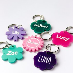 May include: A collection of colorful flower-shaped keychains. Each keychain features a different name in white lettering: Lily, Willow, Lucy, Zoe, Daisy, and Luna. The keychains are in various colors, including purple, green, pink, and teal.