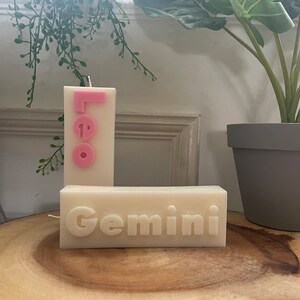 May include: Two cream-colored candles with zodiac signs. One candle reads "Leo" in pink letters, and the other reads "Gemini" in white letters. The candles are on a wooden surface, with greenery in the background.