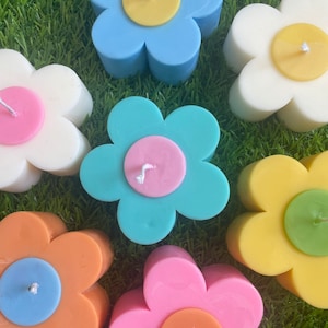 May include: A collection of flower-shaped candles in various pastel colors, including blue, white, pink, orange, and yellow. Each candle has a contrasting center and a wick. The candles are arranged on a green surface, creating a cheerful and decorative display.