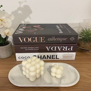 May include: A decorative arrangement featuring a stack of fashion books with the titles "VOGUE", "PRADA", and "COCO CHANEL". Two white bubble candles sit on a marble tray. A potted orchid and a small succulent in a glass container complete the display.