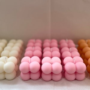 May include: Rows of cube-shaped candles in three colors: ivory, pink, and peach. Each candle has a rounded, bubble-like design and a single wick. The candles are arranged in neat rows, creating a visually appealing display.