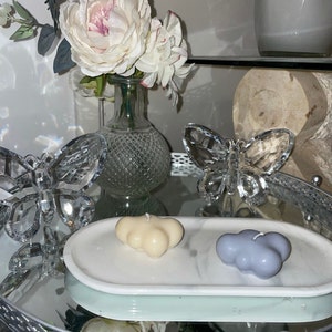 May include: A decorative arrangement featuring cloud-shaped candles in cream and blue hues, resting on a marble-patterned tray. Crystal butterfly figurines and a vase of white flowers add elegance. The tray sits on a mirrored surface.