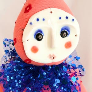 Puede incluir: Mu&ntilde;eca hecha a mano con rostro de cer&aacute;mica y cuerpo de tela rosa. La mu&ntilde;eca tiene un cuello con lentejuelas azules y un sombrero rosa con detalles de labios rojos. La cara tiene maquillaje de ojos azul y mejillas rosadas.