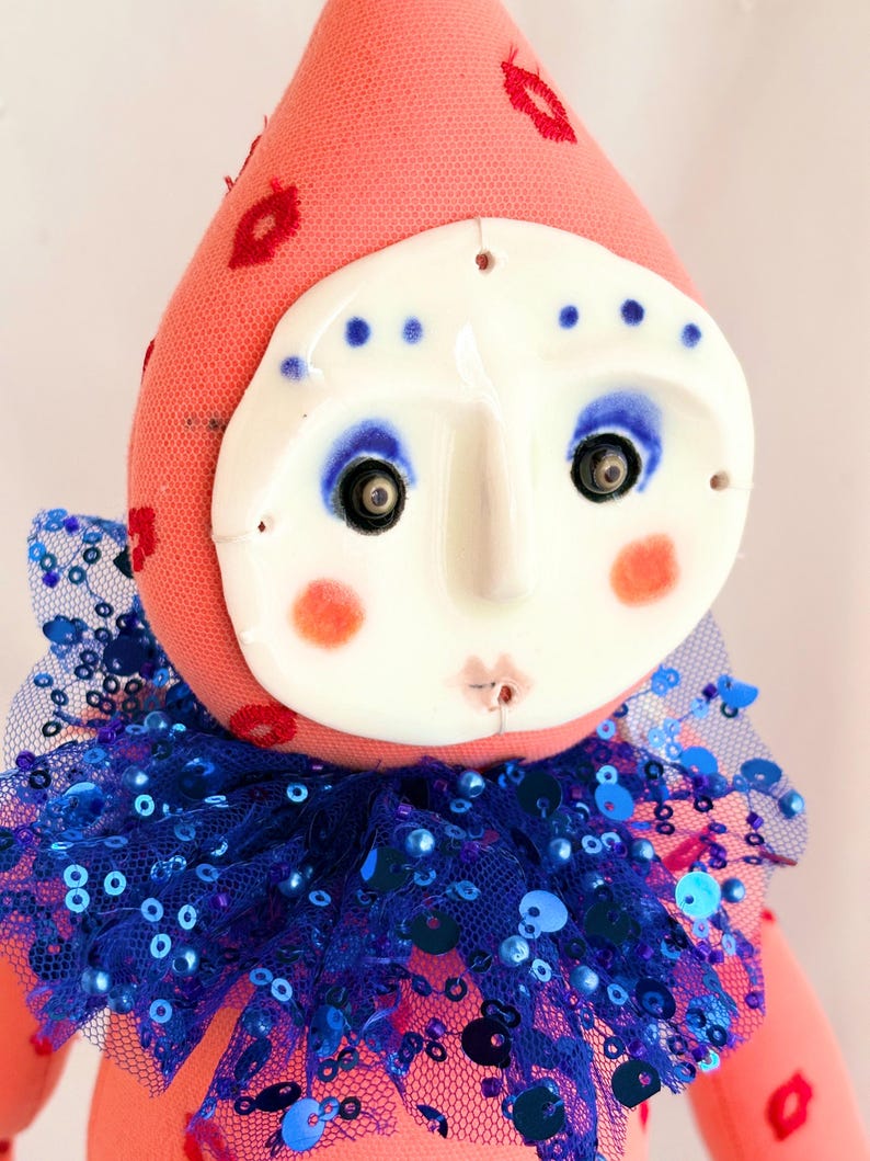 Puede incluir: Mu&ntilde;eca hecha a mano con rostro de cer&aacute;mica y cuerpo de tela rosa. La mu&ntilde;eca tiene un cuello con lentejuelas azules y un sombrero rosa con detalles de labios rojos. La cara tiene maquillaje de ojos azul y mejillas rosadas.