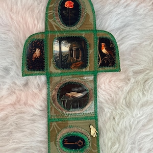 K&ouml;nnte beinhalten: Ein handgefertigtes Stoffkunstwerk mit einem gr&uuml;nen und goldenen Blumenmuster. Es zeigt ovale Tafeln mit Bildern einer Rose, einer Eule, einer Landschaft, eines Rotkehlchens, eines offenen Buches, eines Schl&uuml;ssels und einer kleinen goldenen Hand. Die R&auml;nder sind mit gr&uuml;nem Faden gen&auml;ht.