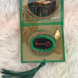 K&ouml;nnte beinhalten: Ein handgefertigter Lesezeichen mit einer gr&uuml;nen Quaste. Das Lesezeichen zeigt zwei kreisf&ouml;rmige Bilder: eines von einem offenen Buch auf einem Tisch und ein weiteres mit einem Schl&uuml;ssel. Das Lesezeichen ist mit gr&uuml;nen N&auml;hten und einem goldfarbenen Handanh&auml;nger verziert.