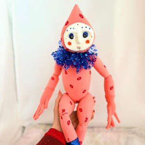Puede incluir: Mu&ntilde;eca hecha a mano con cuerpo rosa y cabeza a juego en forma de l&aacute;grima. La mu&ntilde;eca tiene detalles rojos en forma de labios, un cuello de lentejuelas azul y botas azules. La cara es blanca con ojos azules y mejillas rojas.