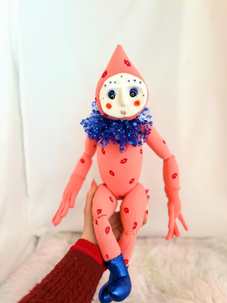 Puede incluir: Mu&ntilde;eca hecha a mano con cuerpo rosa y cabeza a juego en forma de l&aacute;grima. La mu&ntilde;eca tiene detalles rojos en forma de labios, un cuello de lentejuelas azul y botas azules. La cara es blanca con ojos azules y mejillas rojas.