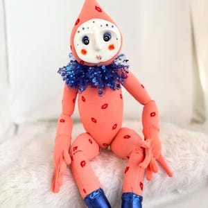Puede incluir: Una mu&ntilde;eca caprichosa con un cuerpo color melocot&oacute;n y una cabeza en forma de l&aacute;grima. La mu&ntilde;eca tiene una cara blanca como una m&aacute;scara con ojos azules y rubor rojo. Marcas de l&aacute;piz labial rojo adornan el cuerpo. Lleva un cuello de lentejuelas azul y botas azules.