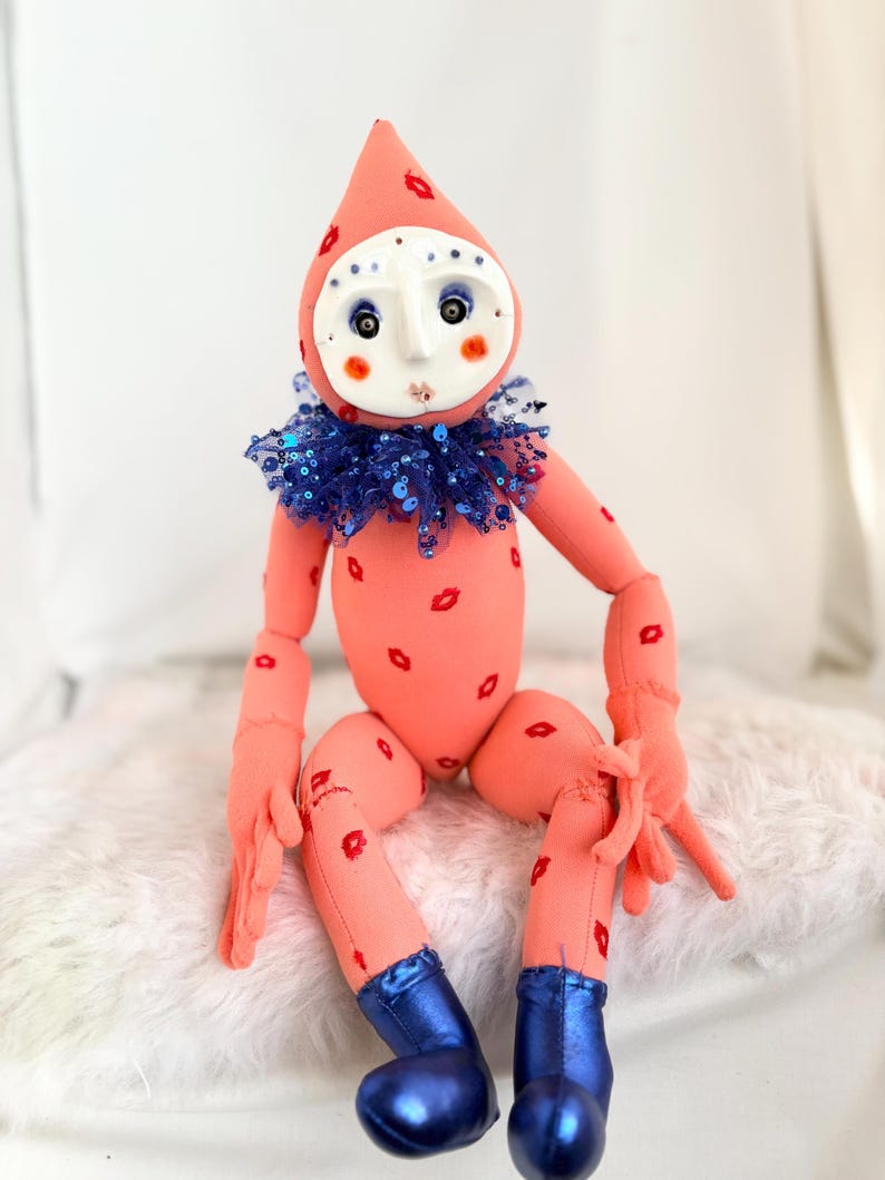 Puede incluir: Una mu&ntilde;eca caprichosa con un cuerpo color melocot&oacute;n y una cabeza en forma de l&aacute;grima. La mu&ntilde;eca tiene una cara blanca como una m&aacute;scara con ojos azules y rubor rojo. Marcas de l&aacute;piz labial rojo adornan el cuerpo. Lleva un cuello de lentejuelas azul y botas azules.