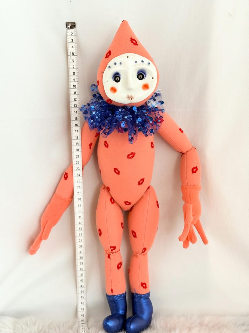Puede incluir: Mu&ntilde;eca hecha a mano con cuerpo color melocot&oacute;n y cabeza en forma de cono. La mu&ntilde;eca tiene una cara de m&aacute;scara blanca, un cuello de lentejuelas azules y detalles rojos en forma de labios. La mu&ntilde;eca mide unos 122 cm de altura.