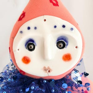 Puede incluir: Mu&ntilde;eca hecha a mano con rostro de cer&aacute;mica y cuerpo de tela. La mu&ntilde;eca tiene una cabeza color melocot&oacute;n con labios rojos bordados y puntos azules. La cara es blanca con maquillaje de ojos azul, mejillas naranjas y una boca peque&ntilde;a. Un cuello de lentejuelas azul completa el look.