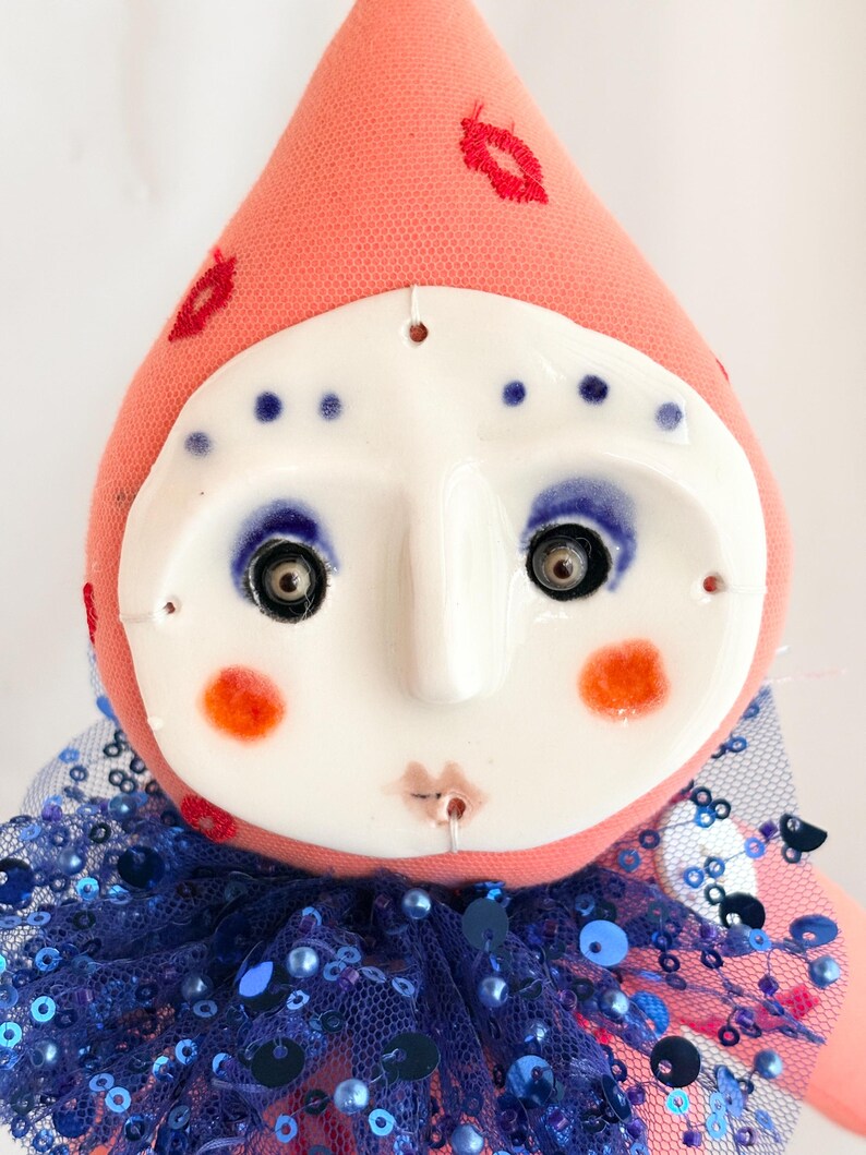 Puede incluir: Mu&ntilde;eca hecha a mano con rostro de cer&aacute;mica y cuerpo de tela. La mu&ntilde;eca tiene una cabeza color melocot&oacute;n con labios rojos bordados y puntos azules. La cara es blanca con maquillaje de ojos azul, mejillas naranjas y una boca peque&ntilde;a. Un cuello de lentejuelas azul completa el look.
