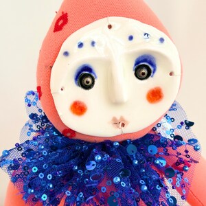 Puede incluir: Mu&ntilde;eca hecha a mano con cuerpo de tela rosa y rostro de cer&aacute;mica blanca. La mu&ntilde;eca tiene ojos azules, mejillas naranjas y un cuello con lentejuelas azules. Bordados rojos con forma de labios decoran el sombrero rosa.