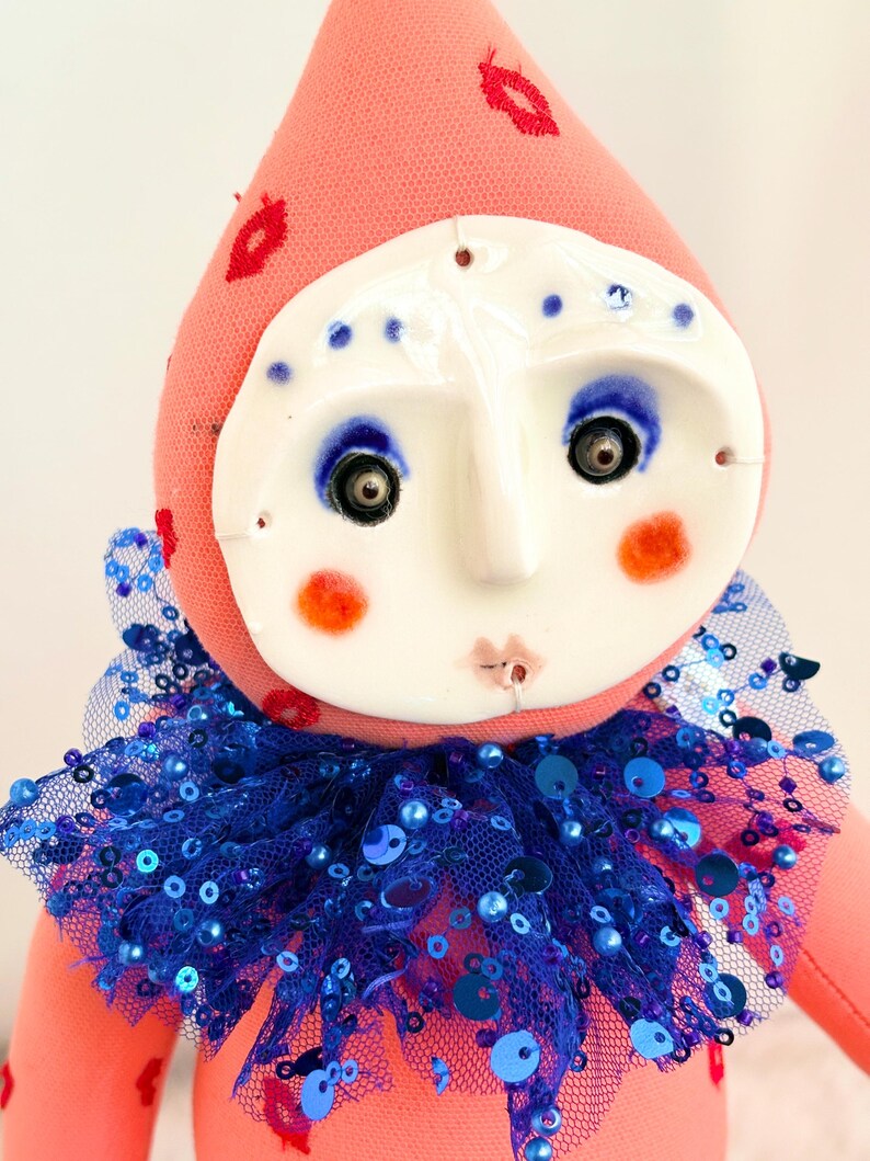 Puede incluir: Mu&ntilde;eca hecha a mano con cuerpo de tela rosa y rostro de cer&aacute;mica blanca. La mu&ntilde;eca tiene ojos azules, mejillas naranjas y un cuello con lentejuelas azules. Bordados rojos con forma de labios decoran el sombrero rosa.