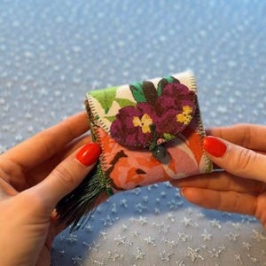 K&ouml;nnte beinhalten: Eine kleine, quadratische florale Geldb&ouml;rse mit Klappenverschluss. Die Geldb&ouml;rse hat ein farbenfrohes Blumenmuster mit lila und gelben Blumen. Eine gr&uuml;ne Quaste h&auml;ngt an der Seite. Die Geldb&ouml;rse wird in zwei H&auml;nden gehalten.