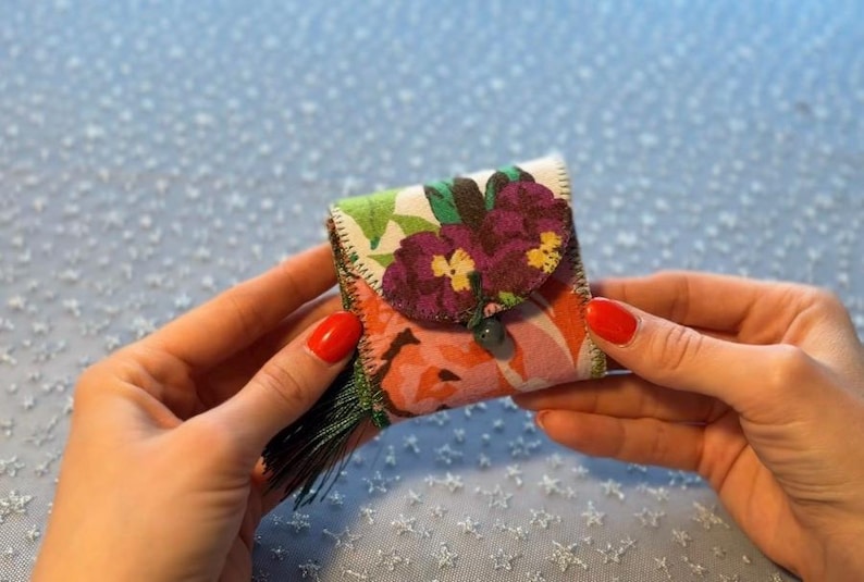 K&ouml;nnte beinhalten: Eine kleine, quadratische florale Geldb&ouml;rse mit Klappenverschluss. Die Geldb&ouml;rse hat ein farbenfrohes Blumenmuster mit lila und gelben Blumen. Eine gr&uuml;ne Quaste h&auml;ngt an der Seite. Die Geldb&ouml;rse wird in zwei H&auml;nden gehalten.