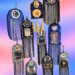 Op de afbeelding: Een collectie van tien hangende wandornamenten in diverse vormen, waaronder ovale en boogvormige ontwerpen. Elk stuk is versierd met gedetailleerd kralenwerk, lovertjes en lange franjes, met vintage-stijl illustraties van manen, ogen en figuren.