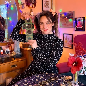 K&ouml;nnte beinhalten: Eine Frau h&auml;lt ein kleines Objekt im Vintage-Stil mit mehreren Tafeln und Illustrationen. Sie tr&auml;gt ein schwarzes Kleid mit wei&szlig;en Sternen. Ein Tisch vor ihr ist mit Tarotkarten und einem gemusterten Tuch bedeckt. Der Raum ist mit Kunst und Lichterketten dekoriert.