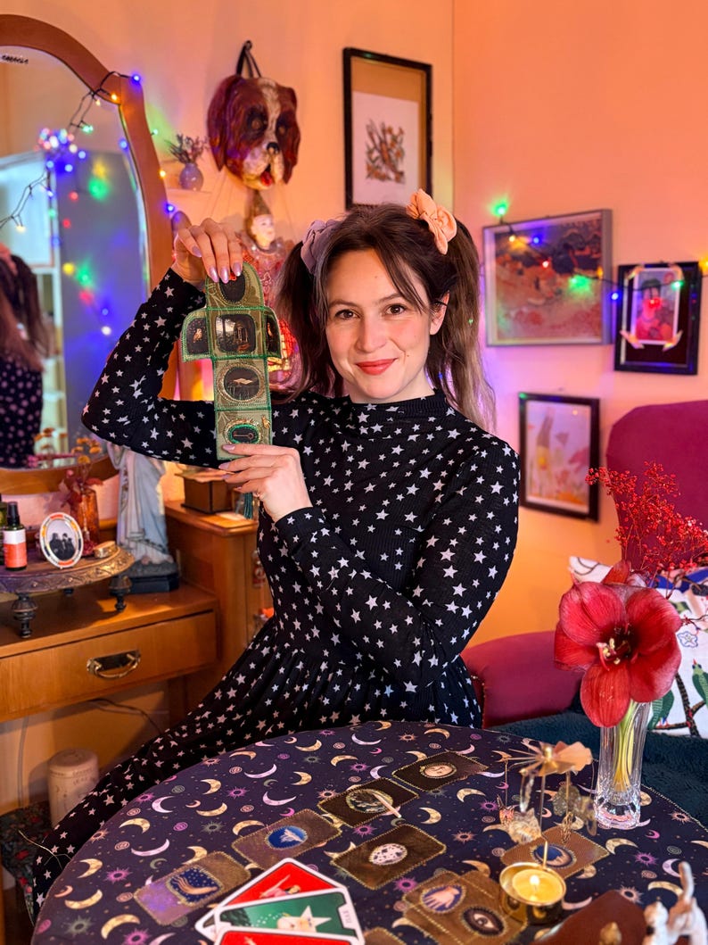 K&ouml;nnte beinhalten: Eine Frau h&auml;lt ein kleines Objekt im Vintage-Stil mit mehreren Tafeln und Illustrationen. Sie tr&auml;gt ein schwarzes Kleid mit wei&szlig;en Sternen. Ein Tisch vor ihr ist mit Tarotkarten und einem gemusterten Tuch bedeckt. Der Raum ist mit Kunst und Lichterketten dekoriert.