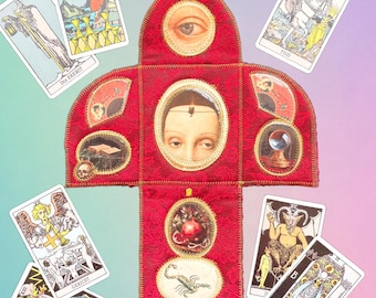 May include: A cross-shaped red fabric textile featuring embroidered edges and multiple circular collaged images, including a human eye, a face, a pomegranate, and a scorpion, surrounded by seven tarot cards with various illustrations.