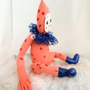 Puede incluir: Una mu&ntilde;eca hecha a mano con un cuerpo color melocot&oacute;n y un gorro puntiagudo adornado con formas de flores rojas. La mu&ntilde;eca tiene una cara blanca en forma de m&aacute;scara, un cuello de lentejuelas azules y botas azules. Formas de labios rojos est&aacute;n dispersas por el cuerpo.
