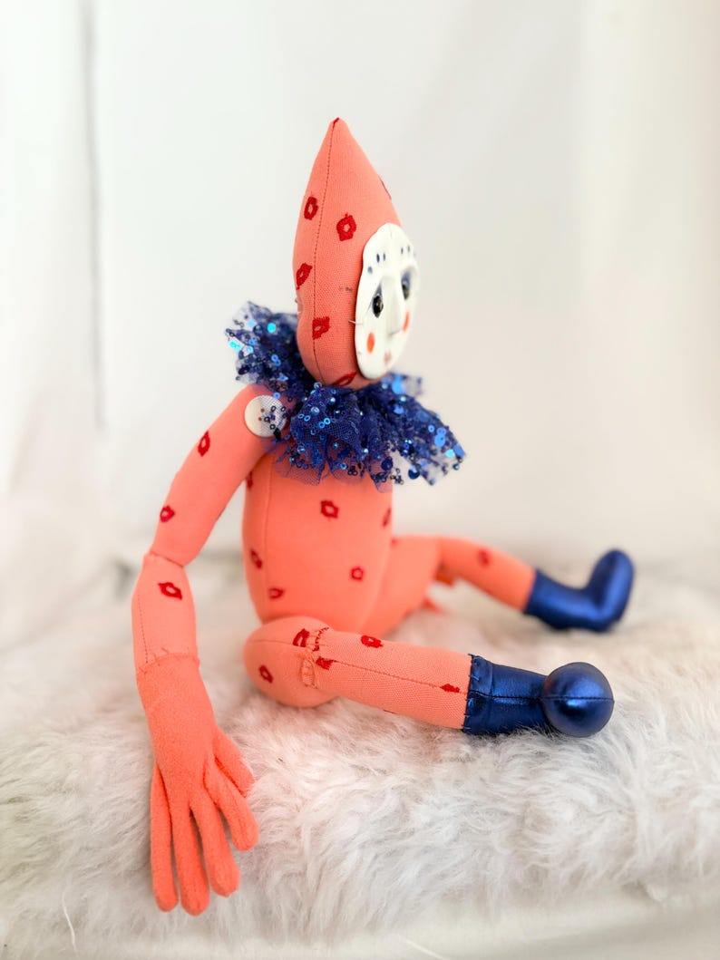 Puede incluir: Una mu&ntilde;eca hecha a mano con un cuerpo color melocot&oacute;n y un gorro puntiagudo adornado con formas de flores rojas. La mu&ntilde;eca tiene una cara blanca en forma de m&aacute;scara, un cuello de lentejuelas azules y botas azules. Formas de labios rojos est&aacute;n dispersas por el cuerpo.