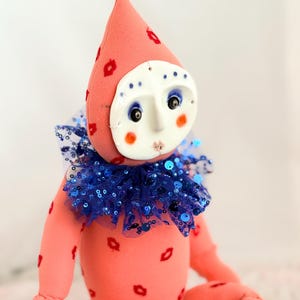 Puede incluir: Mu&ntilde;eca hecha a mano con un dise&ntilde;o caprichoso. La mu&ntilde;eca presenta un cuerpo rosa adornado con huellas de labios rojos, una cara blanca con rasgos pintados y un cuello azul con lentejuelas. El sombrero de la mu&ntilde;eca tambi&eacute;n es rosa con huellas de labios rojos.