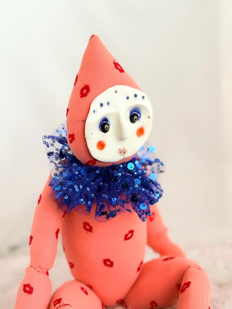 Puede incluir: Mu&ntilde;eca hecha a mano con un dise&ntilde;o caprichoso. La mu&ntilde;eca presenta un cuerpo rosa adornado con huellas de labios rojos, una cara blanca con rasgos pintados y un cuello azul con lentejuelas. El sombrero de la mu&ntilde;eca tambi&eacute;n es rosa con huellas de labios rojos.