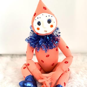 Puede incluir: Mu&ntilde;eca hecha a mano con cuerpo color melocot&oacute;n y un gorro puntiagudo adornado con huellas de labios rojos. La mu&ntilde;eca tiene una cara blanca con ojos azules, mejillas rosadas y un cuello de lentejuelas azules. Lleva botas met&aacute;licas azules.