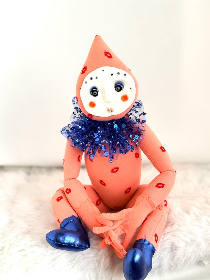 Puede incluir: Mu&ntilde;eca hecha a mano con cuerpo color melocot&oacute;n y un gorro puntiagudo adornado con huellas de labios rojos. La mu&ntilde;eca tiene una cara blanca con ojos azules, mejillas rosadas y un cuello de lentejuelas azules. Lleva botas met&aacute;licas azules.