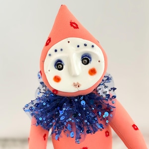 Puede incluir: Mu&ntilde;eca hecha a mano con cuerpo color melocot&oacute;n y sombrero c&oacute;nico adornado con huellas de labios rojos. Presenta una cara de cer&aacute;mica blanca con ojos azules, mejillas rosadas y un cuello con lentejuelas azules. Dise&ntilde;o caprichoso y art&iacute;stico.