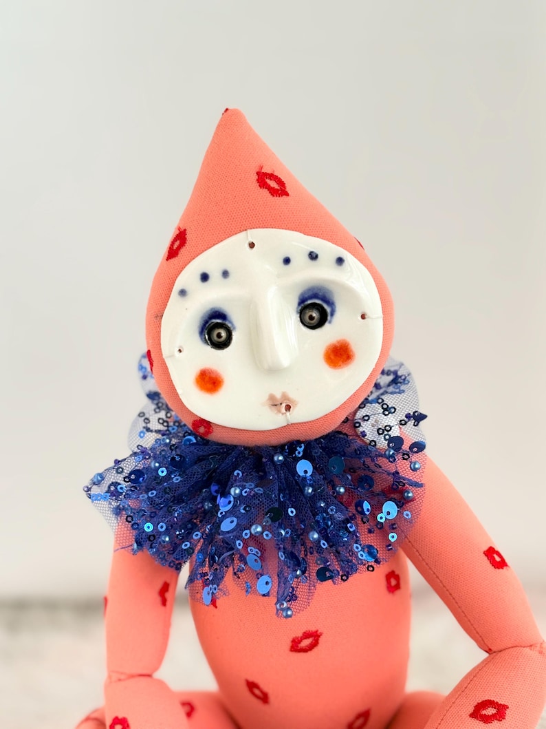 Puede incluir: Mu&ntilde;eca hecha a mano con cuerpo color melocot&oacute;n y sombrero c&oacute;nico adornado con huellas de labios rojos. Presenta una cara de cer&aacute;mica blanca con ojos azules, mejillas rosadas y un cuello con lentejuelas azules. Dise&ntilde;o caprichoso y art&iacute;stico.