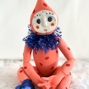 Puede incluir: Una mu&ntilde;eca caprichosa con un cuerpo color melocot&oacute;n adornado con huellas de labios rojos. Presenta una cabeza en forma de l&aacute;grima, una cara blanca en forma de m&aacute;scara con ojos azules y un cuello con lentejuelas azules. La mu&ntilde;eca lleva zapatos met&aacute;licos azules.